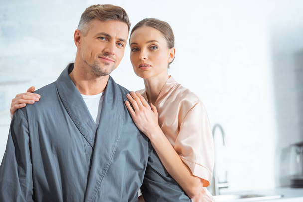 beautiful woman in robe looking at camera and embracing man in kitchen - Photo, Image