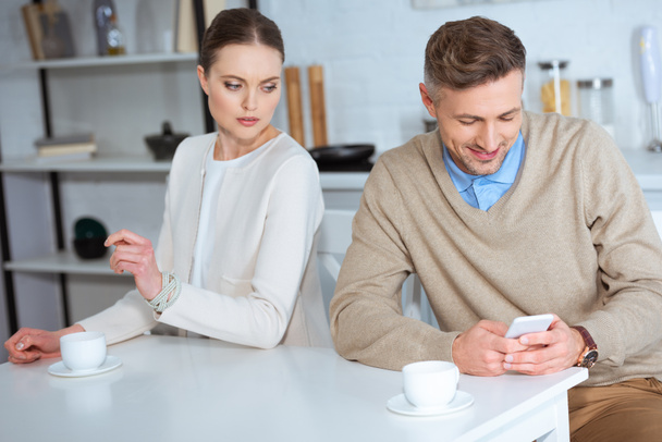 smiling man using smartphone and ignoring woman during breakfast in kitchen - Photo, Image