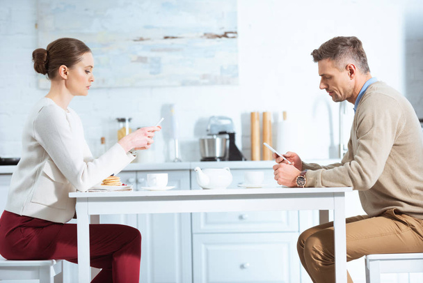 casal adulto usando smartphones e ignorando uns aos outros durante o café da manhã na cozinha
 - Foto, Imagem