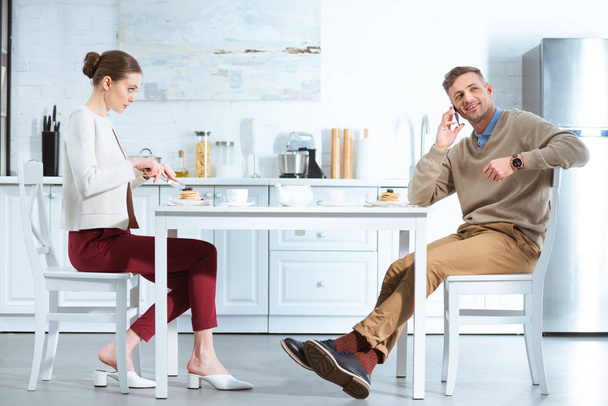 τηγανίτες κοπής δυσαρεστημένοι γυναίκα ενώ ο άνθρωπος μιλά στο smartphone κατά τη διάρκεια του πρωινού στην κουζίνα - Φωτογραφία, εικόνα