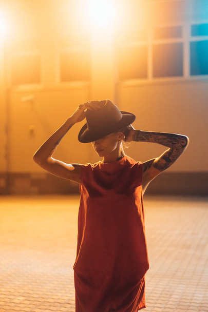 The young, attractive girl in a hat poses to the camera at night - Foto, Imagen