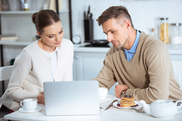 adult couple sitting at table and using laptop during breakfast in morning - Photo, Image