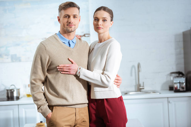 adult couple in casual clothes looking at camera and embracing in kitchen - Photo, image