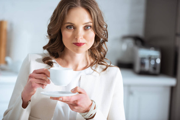 beautiful woman holding coffee cup and looking at camera during breakfast in kitchen - Photo, Image