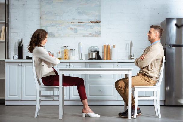 dissatisfied couple looking at each other while sitting with arms crossed at table in kitchen - Foto, immagini