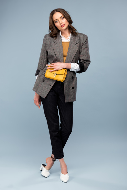 beautiful stylish woman in formal wear holding bag and posing on grey background - 写真・画像