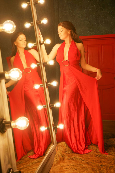 encantadora chica hermosa en un vestido de noche largo rojo en una alfombra de piel cerca de un espejo enorme en un marco con bombillas
 - Foto, imagen