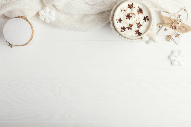 A cup of hot winter drink with whipped cream and a dusting powder with an asterisk, white snowflakes and knitted scarf on a wooden table. Christmas concept in bright colors. Copy space. - Photo, image