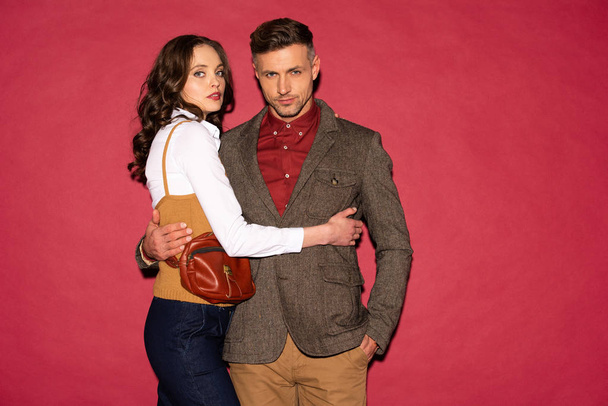 beautiful fashionable couple in formal wear embracing and posing on red background - Zdjęcie, obraz