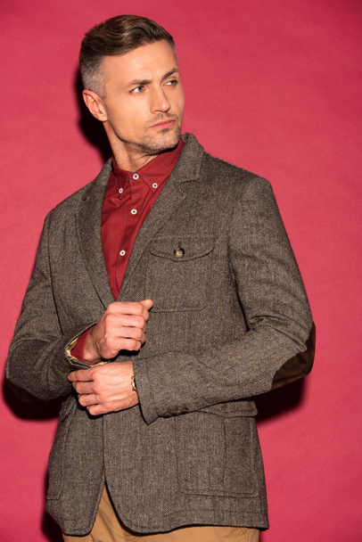 homem bonito em desgaste formal olhando para longe ao ajustar jaqueta no fundo vermelho
 - Foto, Imagem