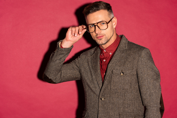 stylischer Mann in formaler Kleidung, der in die Kamera blickt und die Brille auf rotem Hintergrund justiert - Foto, Bild