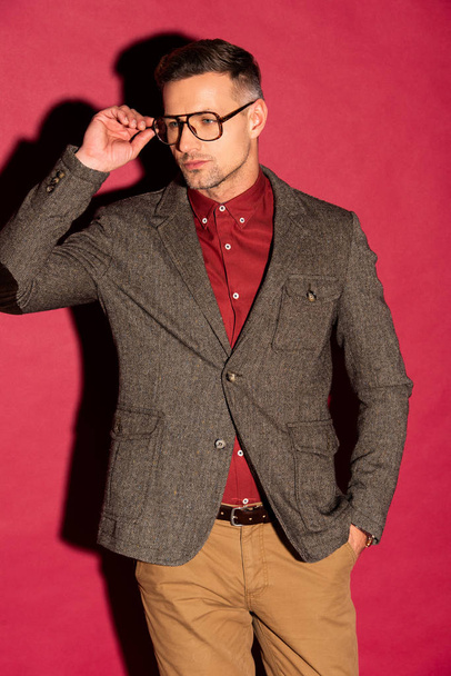 confident stylish man in formal wear and glasses looking away on red background - Фото, зображення