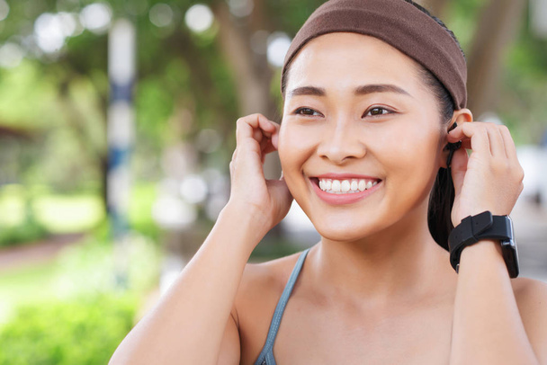 Mladý šťastný asijské žena nošení čelenku a uvedení do sluchátek při práci v parku  - Fotografie, Obrázek