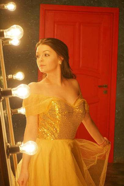 beautiful young girl in a gold evening dress stands on a fur rug near a large mirror in a frame with lights and looks into her reflection - Photo, Image