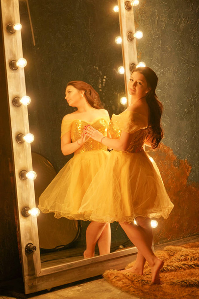 schöne junge Mädchen in einem goldenen Abendkleid steht auf einem Pelzteppich neben einem großen Spiegel in einem Rahmen mit Lichtern und blickt in ihr Spiegelbild - Foto, Bild