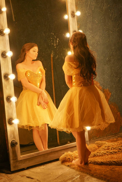 belle jeune fille dans une robe de soirée en or se tient sur un tapis de fourrure près d'un grand miroir dans un cadre avec des lumières et regarde dans son reflet
 - Photo, image