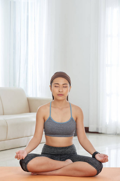 Concentrated Asian sportswoman sitting on mat at home and meditating with eyes closed in soft daylight - Fotografie, Obrázek