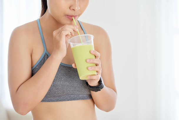 Crop shot of sportive woman drinking green smoothie with straw standing indoors - Фото, изображение