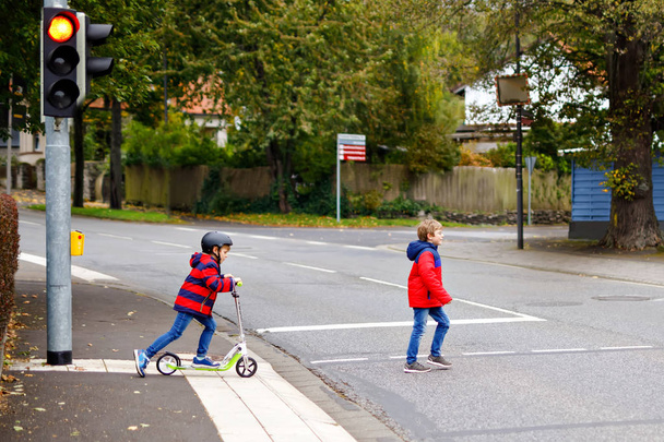 Two little schoolkids boys running and driving on scooter on autumn day. Happy children in colorful clothes and city traffic crossing pedestrian crosswalk with traffic lights. - Photo, Image