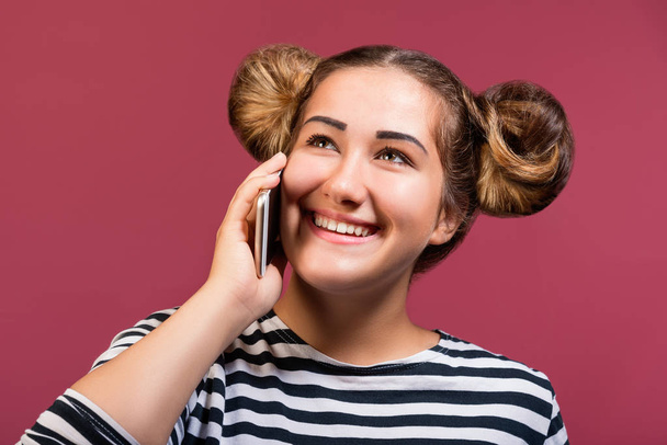 pretty young hipster girl with funny hairstyle and in striped shirt talking on the phone with smiling face, isolated over pink background - Foto, afbeelding