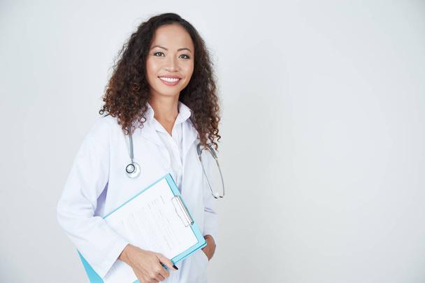 Porträt einer Ärztin mit lockigem Haar im weißen Mantel, die mit Gesundheitskarte steht und lächelt - Foto, Bild