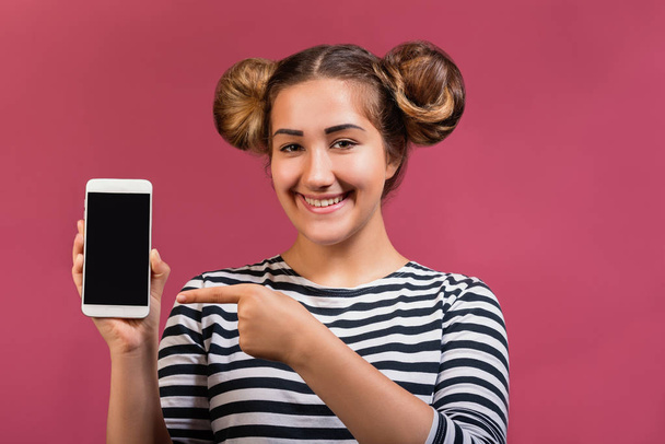 Jovem feliz hipster menina com penteado engraçado apontando dedo para tela em branco telefone celular isolado sobre fundo rosa
 - Foto, Imagem