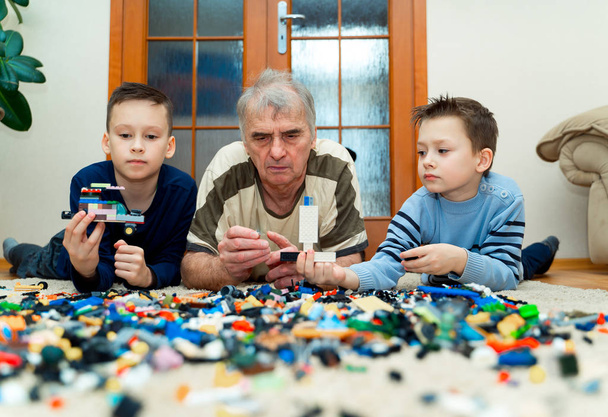 Grandfather with his grandsons lying on the floor and building with lego bricks at home on the background of the glass door. Old and new generation - Fotoğraf, Görsel