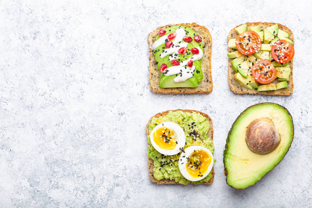 Avocado toasts with egg, tomatoes, seasonings and a half of whole avocado over white stone background, space for text. Healthy breakfast avocado sandwiches with different toppings, top view, close-u - Foto, afbeelding