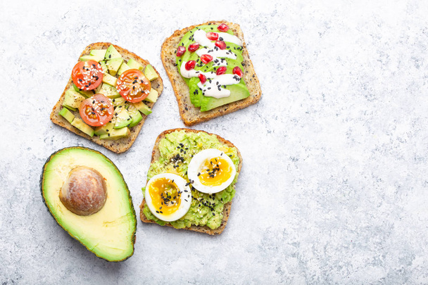 Avocado toasts with egg, tomatoes, seasonings and a half of whole avocado over white stone background, space for text. Healthy breakfast avocado sandwiches with different toppings, top view, close-u - Photo, Image