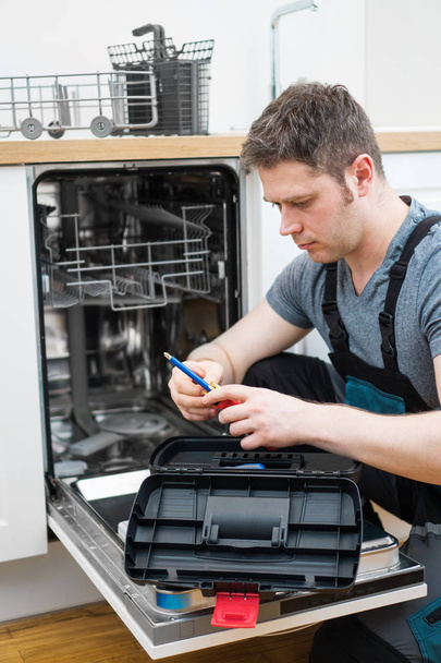 Professional handyman in overalls repairing domestic dishwasher in the kitchen. - Photo, Image