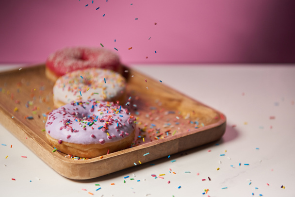 selective focus of tasty donuts with icing and falling sprinkles on wooden cutting board  - Fotó, kép