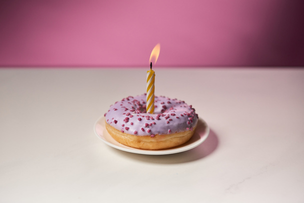 burning candle in middle of doughnut with sprinkles on white table on pink background - Foto, imagen