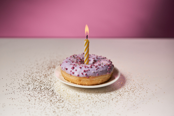 brandende kaars in midden van geglazuurde donut met sparkles op witte tafel op roze achtergrond - Foto, afbeelding
