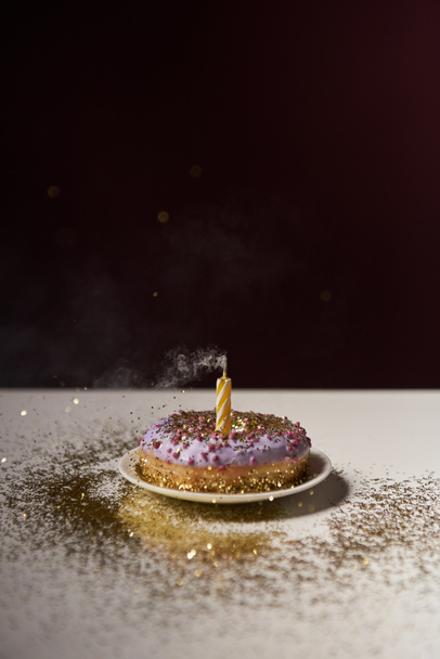 candle in middle of doughnut with golden sparkles on white table isolated on black - Foto, imagen