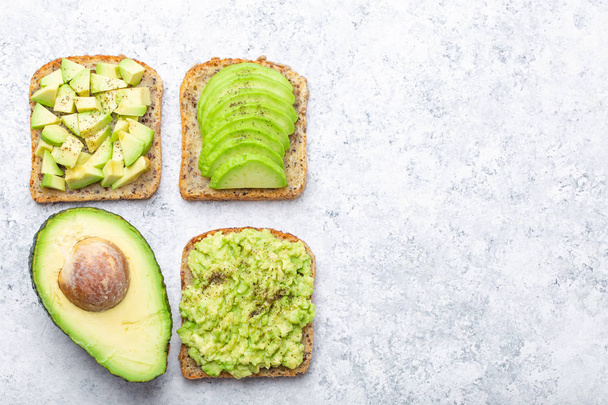 Different types of avocado toasts and a half of whole avocado over white stone background, space for text. Ways of making and serving delicious healthy breakfast avocado sandwich, top view, close-u - Photo, Image