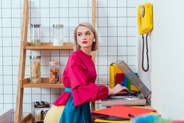 beautiful housewife in colorful clothes listening music on vinyl record player and posing in kitchen - Photo, Image