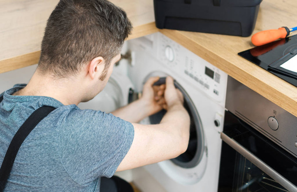 Professional handyman in overalls repairing washing machine in the kitchen. - Fotó, kép
