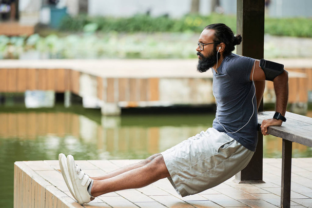 Vista lateral del hombre indio barbudo haciendo ejercicio de calentamiento al aire libre y escuchando música con auriculares
 - Foto, imagen