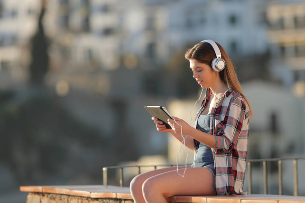 Side view portrait of a serious teenage girl e-learning using a tablet sitting on a ledge in a town - 写真・画像