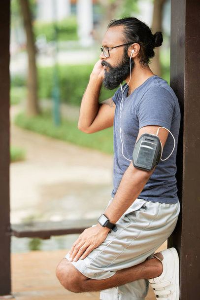 Вид сбоку на бородатого индийца в спортивной одежде со смартфоном на плече, стоящего на открытом воздухе, отдыхающего после тренировки
  - Фото, изображение