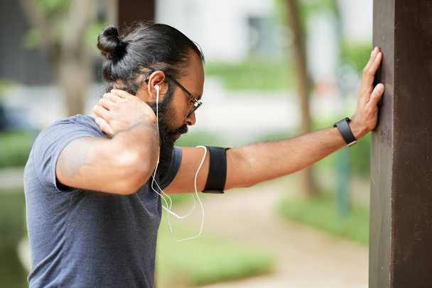 Boční pohled na sportovce s tmavovlasým vousem, který zahřívá venkovní cvičení a poslouchá hudbu prostřednictvím sluchátek - Fotografie, Obrázek