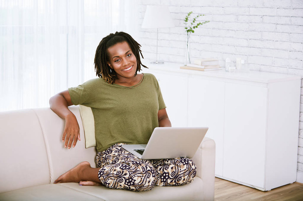 Улыбающаяся афроамериканка в повседневной одежде, работающая дома на ноутбуке
 - Фото, изображение