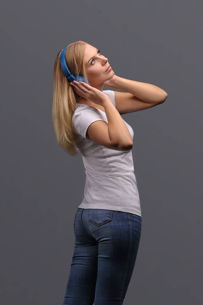 Young girl blonde in blue headphones. Listening, enjoying music. Isolate on a gray background. - Фото, изображение