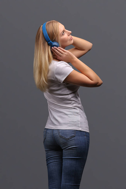 Young girl blonde in blue headphones. Listening, enjoying music. Isolate on a gray background. - 写真・画像