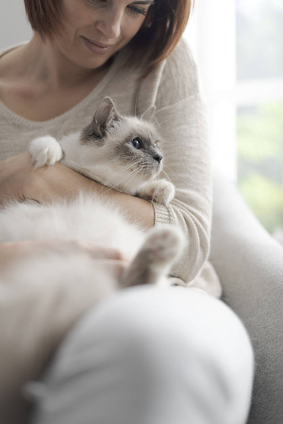 Young happy woman sitting on the armchair in the living room and petting her beautiful cat - Fotografie, Obrázek