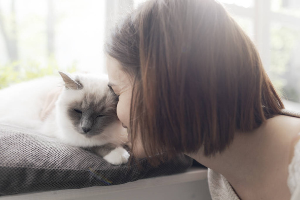 Young woman petting her beautiful cat at home next to a window, pets and lifestyle concept - Foto, afbeelding