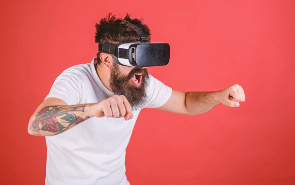 Hipster on excited face driving bike on high speed in virtual reality with modern digital gadget. Man with beard in VR glasses driving motorbike, red background. Virtual driving lessons concept - Photo, Image