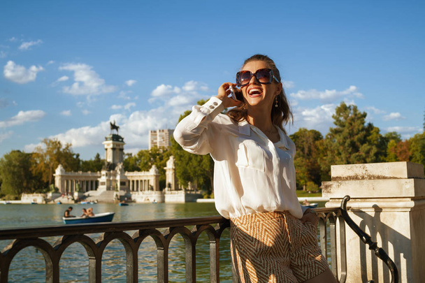 smiling modern woman in white blouse and shorts at Parque del Buen Retiro in Madrid, Spain talking on a cell phone. big fashionable sunglasses. woman talking to family or loved one & share experience. - Photo, image