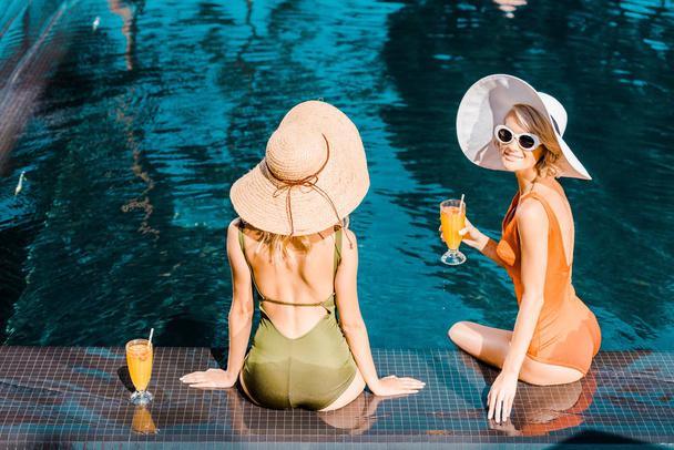 back view of pin up girls in swimsuits and hats sitting at poolside with orange cocktails - Фото, зображення