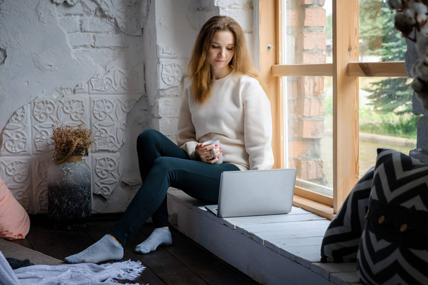 Retrato de una hermosa joven que se sienta por la mañana en el alféizar de la ventana con un libro para ordenador portátil y bebe café
. - Foto, imagen